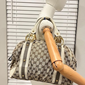 Puede incluir: Un bolso de mano de lona con monograma en beige y marrón que presenta ribetes de cuero blanco, detalles verticales de cuero gris con tachuelas doradas y herrajes de metal dorado con el logotipo GG entrelazado en las asas circulares.
