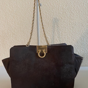 May include: A dark brown handbag with a gold-tone chain strap and a gold-tone clasp. The bag has a rectangular shape with a flap closure and side pockets. The bag is made of a suede-like material.