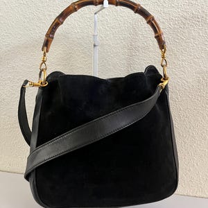 May include: Black suede handbag with a bamboo handle and a detachable black leather shoulder strap. The bag features gold-toned hardware and a soft, unstructured shape. The handle is made of curved, polished bamboo.