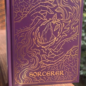 Puede incluir: Un diario morado con detalles dorados. La portada presenta un diseño intrincado de manos sosteniendo cristales, con la palabra "SORCERER" en la parte inferior. Una cinta dorada como marcapáginas es visible. Un dado con el número 20 también es visible.