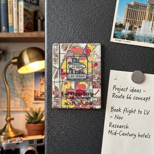 May include: A rectangular magnet featuring a map of the Las Vegas Strip in red, yellow, and grey. The magnet includes the text "Las Vegas Strip" and coordinates. A postcard of the Bellagio fountains is also visible.