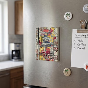 May include: A rectangular magnet featuring a map of the Las Vegas Strip, with red, yellow, and blue design elements. The magnet has the text "Las Vegas Strip" and coordinates. It's attached to a stainless steel fridge door.
