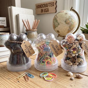 Puede incluir: Tres frascos de plástico transparente con forma de pata y tapas de rosca. El primero contiene pelaje oscuro y una etiqueta que dice PET KEEPSAKE FUR. El segundo contiene clips de colores y una etiqueta que dice DESK ORGANIZER. El tercero contiene pequeñas conchas marinas y una etiqueta que dice TINY TREASURES.