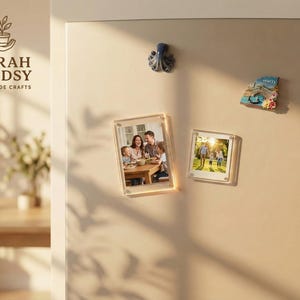 Puede incluir: Una puerta de nevera crema con dos marcos de fotos de acrílico transparente, cada uno con una fotografía familiar. También hay un imán de pulpo azul y un imán de recuerdo. Los marcos son rectangulares. La marca "FARAH LINDSY HANDMADE CRAFTS" es visible.