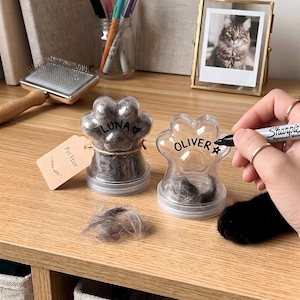 Puede incluir: Dos recipientes de plástico transparente con forma de pata rellenos de pelo de mascota gris. Uno está marcado como LUNA con un corazón, y el otro se está escribiendo con un marcador negro para decir OLIVER con una estrella. Tiene una etiqueta Pet Hair.