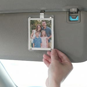 Peut inclure: Un cadre photo en acrylique transparent avec un portrait de famille, fixé à un pare-soleil de voiture. Le cadre est maintenu en place par un clip argenté. La famille sur la photo se compose de deux adultes et de deux enfants.