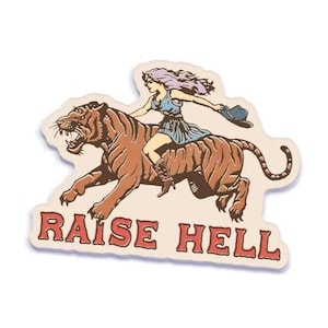 May include: A sticker featuring a woman riding a tiger, with the words "RAISE HELL" in red. The woman has purple hair and wears a blue dress and cowboy boots. The tiger is brown with black stripes.