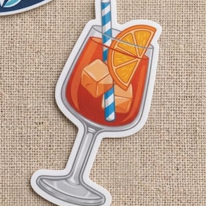 May include: A sticker featuring a cartoon illustration of a cocktail in a stemmed glass. The drink is orange-red with ice cubes, a blue and white striped straw, and an orange slice. The sticker has a white border.