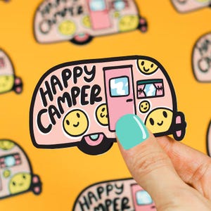 May include: A pink camper sticker with the words "Happy Camper" in black. The sticker has yellow smiley faces and a turquoise door. The background is a repeating pattern of the same camper design.