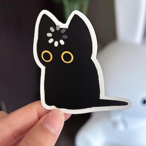 May include: A black cat sticker with yellow eyes and a white border. The cat has a loading symbol on its head. The sticker is held by a hand, with a blurred white object in the background.