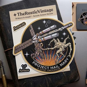 May include: A sticker featuring a space-themed design with the text "Project Hail Mary" and an astronaut. The sticker is on a leather-bound journal with a pen and pencil. A Kindle e-reader with a matching sticker is also visible.