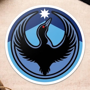 May include: A round sticker featuring a stylized black bird with outstretched wings, a red eye, and a white star. The bird is set against a blue and light blue background with a mountain-like shape.