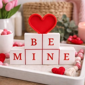 May include: A Valentine's Day themed display on a white tray. It features a red heart, blocks spelling "BE MINE", a cup of pink and white candies, pink tulips, and a lit candle. Decorative beads and hearts are also present.