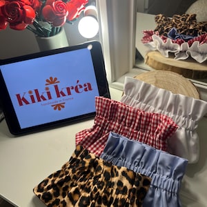 Può includere: Una collezione di elastici per capelli in tessuto con vari motivi e colori, tra cui stampa leopardata, quadretti rossi e bianchi, e blu e bianco tinta unita. Gli elastici sono esposti su una superficie bianca con il logo "Kiki Kréa" su un tablet.