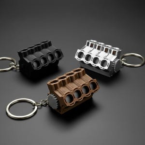 May include: Three miniature engine keychains in black, silver, and brown. Each keychain features a detailed engine block design with a silver keyring attached. The keychains are displayed on a dark gray surface.
