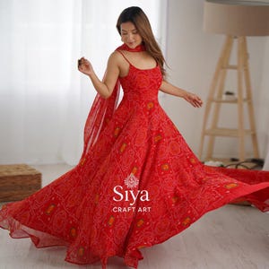 May include: A flowing red dress with a matching scarf. The dress features a detailed pattern in white and orange, with thin straps and a wide skirt. The dress has the text "Siya Craft Art" in white.