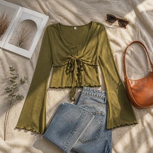 May include: An olive green long-sleeve top with a V-neckline, a front tie detail, and flared sleeves with lettuce-edge hems. The garment is paired with blue denim jeans, a brown leather shoulder bag, and brown tortoise-shell sunglasses.