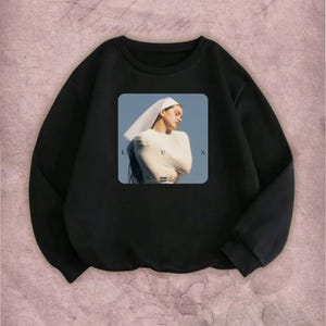 May include: Black sweatshirt featuring a square graphic of a person wearing a white top and head covering, with the letters "L", "U", and "X" visible. The person is set against a blue background.