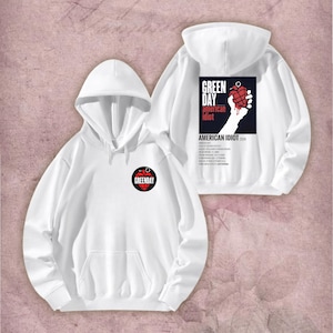 May include: White hoodie featuring a front pocket, hood, and a small circular logo with the text "Green Day". The back of the hoodie displays a graphic with the words "Green Day American Idiot" and a hand holding a heart-shaped grenade.