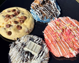 May include: A black plate holds four large, decorated cookies. One is yellow with chocolate chips, another blue with white drizzle and cookie crumbles, a third pink with white drizzle and nuts, and the last with white drizzle, cookie crumbles, and a white chocolate bar.