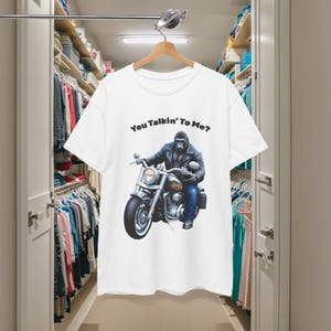 K&ouml;nnte beinhalten: Wei&szlig;es T-Shirt mit einem Gorilla auf einem Motorrad. Der Gorilla tr&auml;gt eine Lederjacke und Jeans. Der Text "You Talkin' To Me?" ist &uuml;ber dem Motiv aufgedruckt. Das T-Shirt h&auml;ngt an einem Holzb&uuml;gel.