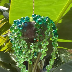 Pode incluir: Um vaso suspenso com um vaso marrom escuro é adornado com fios de contas verdes facetadas. As contas caem em cascata para baixo, criando um efeito decorativo. Uma corrente dourada suspende o vaso contra um fundo de folhas verdes grandes.