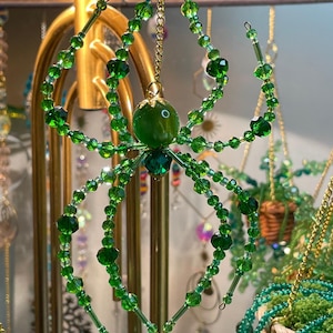 May include: A green beaded spider decoration. The spider has a round green bead body and eight legs made of smaller green beads. It hangs from a gold chain. Ideal for Halloween or as a unique, year-round decorative item.
