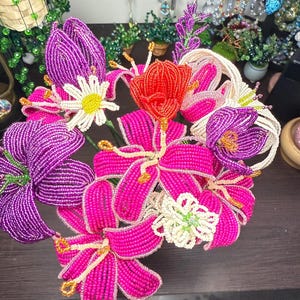May include: A collection of beaded flowers in pink, purple, red, and white. The arrangement features lilies, daisies, and tulips, all made from tiny beads. The flowers are displayed on a dark surface, highlighting the vibrant colours and intricate details of the handcrafted pieces.
