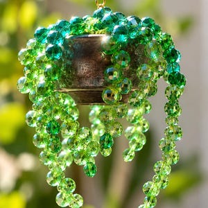 Op de afbeelding: Een hangend ornament met een donker metalen pot gevuld met trapsgewijze strengen van gefacetteerde groene kristallen kralen. De kralen variëren in tinten smaragd- en limoengroen en reflecteren het licht. Een goudkleurige ketting is aan de bovenkant bevestigd.