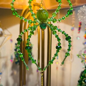 May include: A green beaded spider ornament with a faceted crystal pendant. The spider has a round green body and eight legs made of green beads. The ornament hangs from a gold chain.