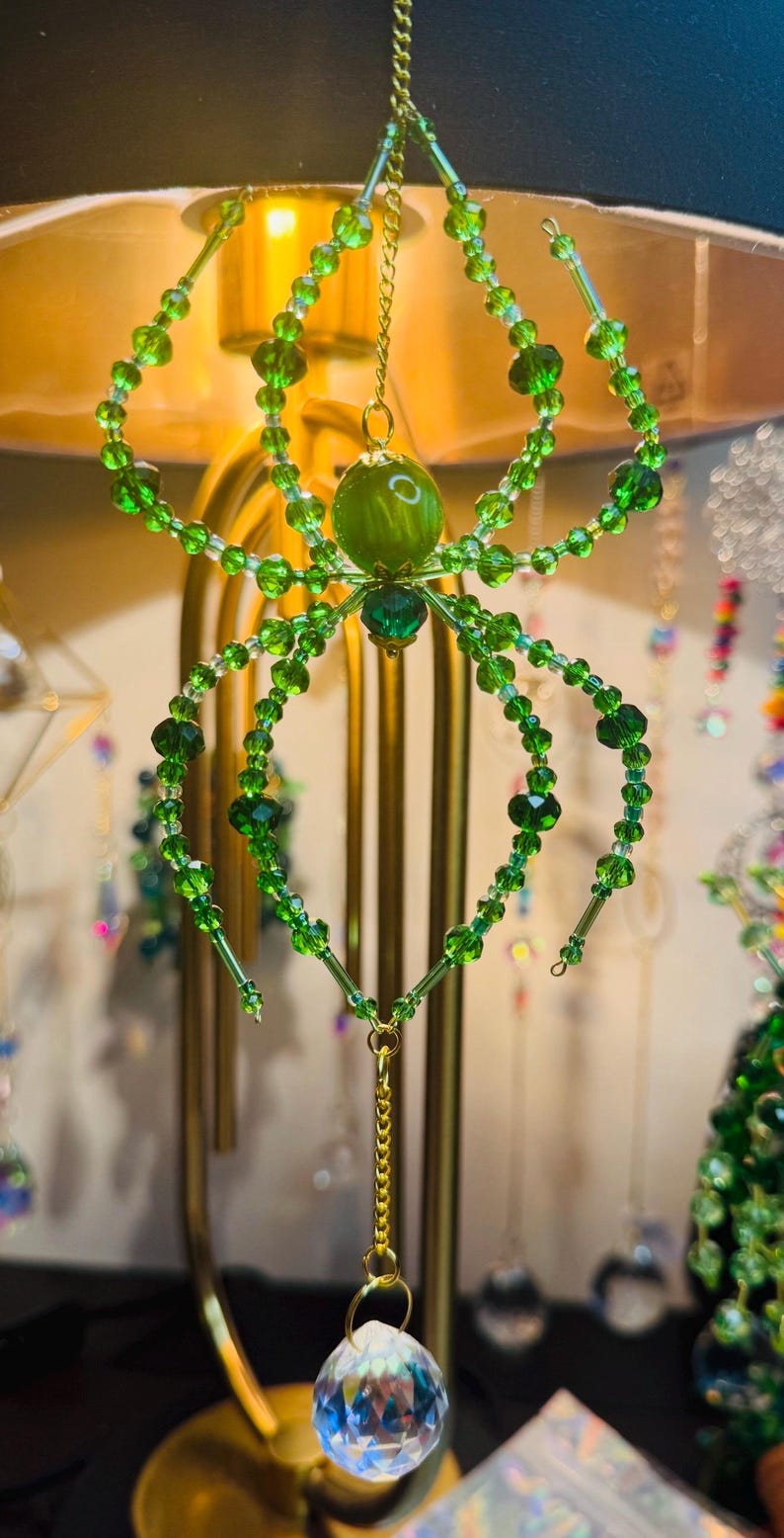 May include: A green beaded spider ornament with a faceted crystal pendant. The spider has a round green body and eight legs made of green beads. The ornament hangs from a gold chain.