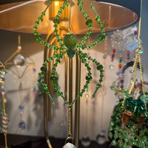 May include: A green beaded spider ornament with a crystal ball pendant. The spider has eight legs and a round body, suspended by a gold chain. Other decorative hanging ornaments are in the background.