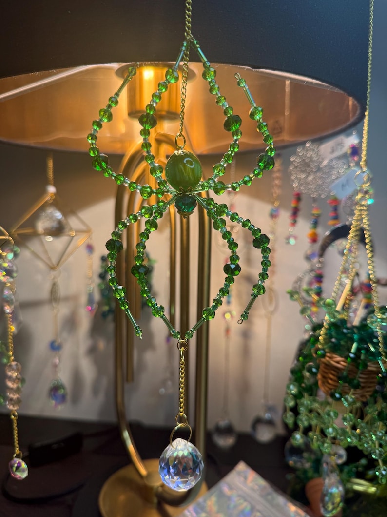 May include: A green beaded spider ornament with a crystal ball pendant. The spider has eight legs and a round body, suspended by a gold chain. Other decorative hanging ornaments are in the background.