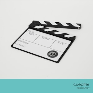 Może przedstawiać: Czarno-biała klaps filmowy z tekstem "PROD., ROLL, SCENE, TAKE, DIRECTOR:, CAMERA:" i okrągłym logo. Klaps jest na białym tle. Jest to narzędzie do tworzenia filmów.