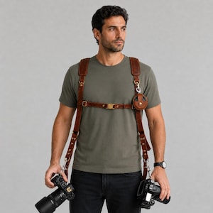 Peut inclure: Un harnais double pour appareil photo en cuir marron avec des sangles réglables, des boucles en métal doré et une petite pochette circulaire fixée sur le côté gauche. Le harnais est porté sur un t-shirt vert olive et un pantalon noir.