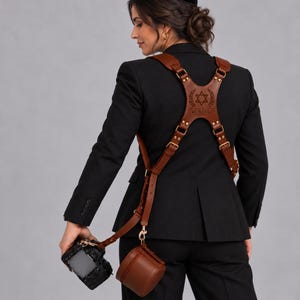 May include: A brown leather dual camera harness featuring adjustable straps, gold-toned metal buckles, and a central back panel engraved with a Star of David and the text YOUR LOGO. The harness includes a small matching leather pouch and clips.
