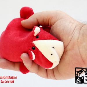 May include: A red and white plush toy in the shape of a small animal. The toy has black eyes and a white nose. The toy is being held in a person's hand. Downloadable PDF tutorial.
