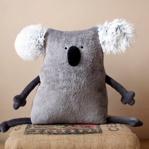 May include: A gray plush koala bear with white fluffy ears, black eyes, and a black nose. The koala has dark gray arms and legs, and is sitting on a burlap sack. The koala is a stuffed animal.