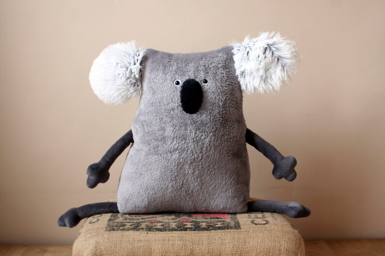 May include: A gray plush koala bear with white fluffy ears, black eyes, and a black nose. The koala has dark gray arms and legs, and is sitting on a burlap sack. The koala is a stuffed animal.