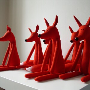May include: Five red fabric fox plushies with long limbs and tails. The foxes are arranged in a row on a white surface.