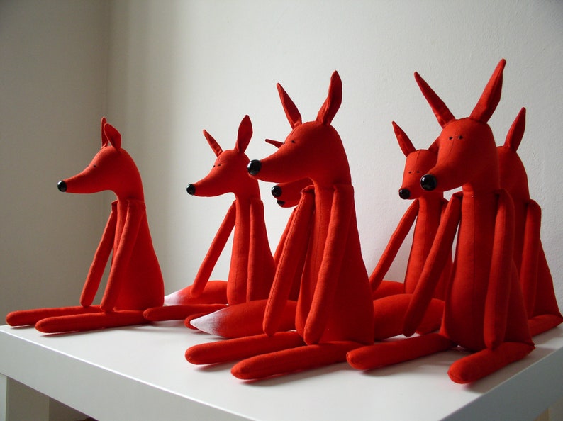 May include: Five red fabric fox plushies with long limbs and tails. The foxes are arranged in a row on a white surface.