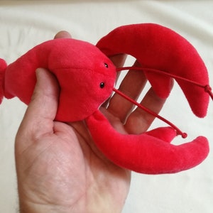 May include: A red plush lobster toy with black eyes. The lobster is held in a person's hand.