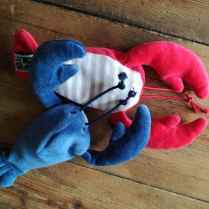May include: A plush red and blue lobster toy with white and blue stripes on its body. The lobster has two large claws and a long tail.