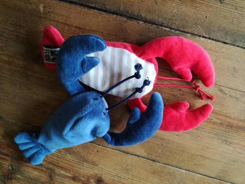 May include: A plush red and blue lobster toy with white and blue stripes on its body. The lobster has two large claws and a long tail.