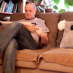 May include: A plush gray koala pillow with white ear accents sits on a brown couch next to a person reading a newspaper. The person is wearing a striped shirt and jeans. The newspaper has the word "thon" visible.