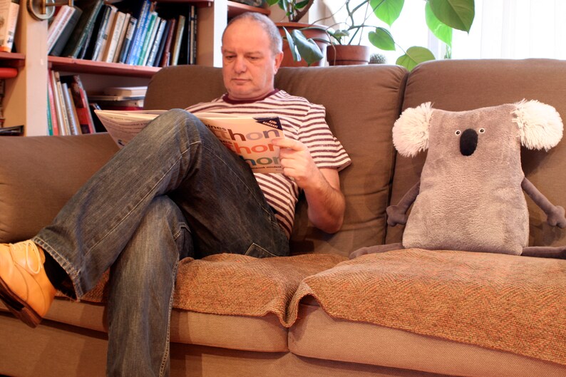 May include: A plush gray koala pillow with white ear accents sits on a brown couch next to a person reading a newspaper. The person is wearing a striped shirt and jeans. The newspaper has the word "thon" visible.