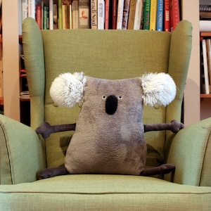 May include: A plush koala bear pillow with a gray body, white fluffy ears, and a black nose and eyes. The koala is sitting on a green armchair in front of a bookshelf. The koala's arms and legs are outstretched.