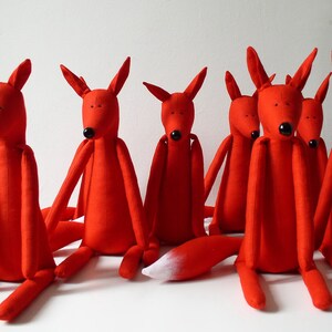 May include: A group of seven red fox plush toys sitting on a white surface. The foxes have long, thin limbs and black eyes. One fox has a white tail tip.