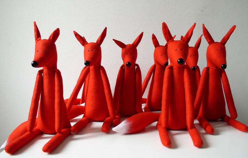 May include: A group of seven red fox plush toys sitting on a white surface. The foxes have long, thin limbs and black eyes. One fox has a white tail tip.