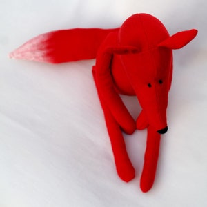 May include: A red plush fox toy with a white tipped tail. The fox is sitting on a white surface.